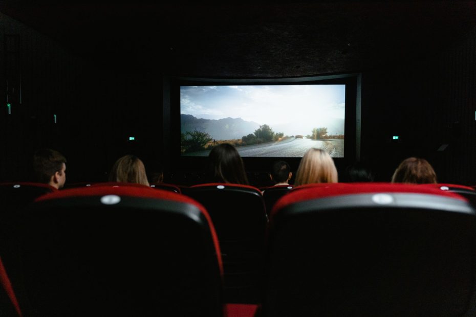 View from behind of people watching a movie in a cinema with red seats and a large screen.